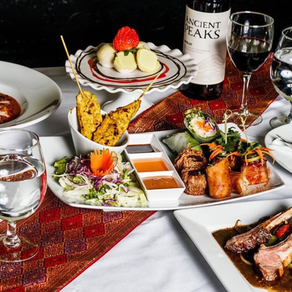 A table laden with various Thai dishes, including satay, spring rolls, salad, and lamb chops.